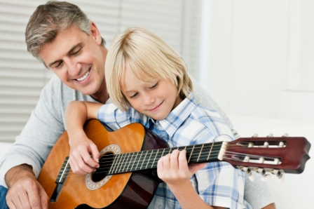 Father and son sit together while the son plays guitar under his father's supervision. Horizontal shot.